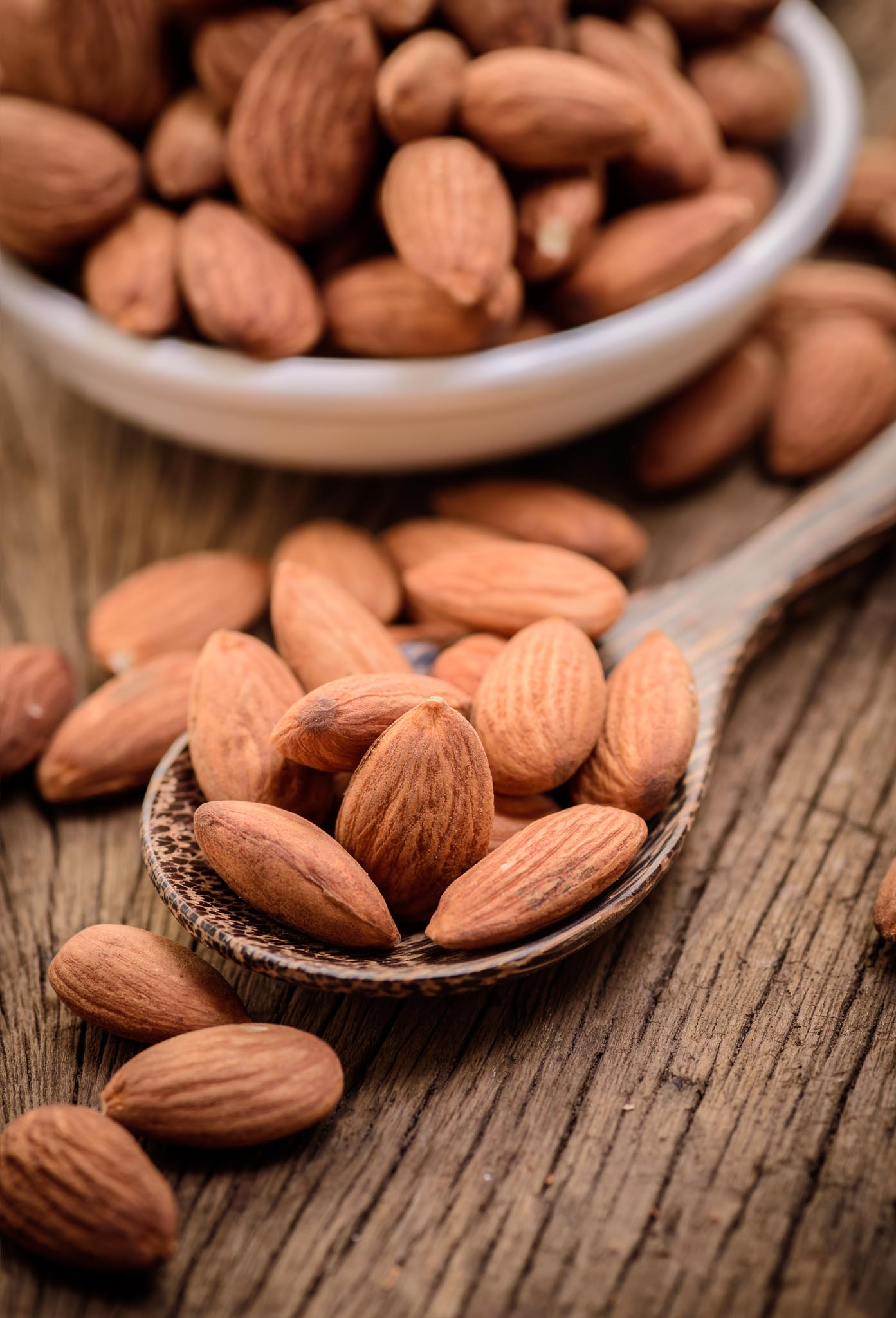 Sweet apricot kernels on a wooden spoon and a white bowl filled with kernels in the background. 