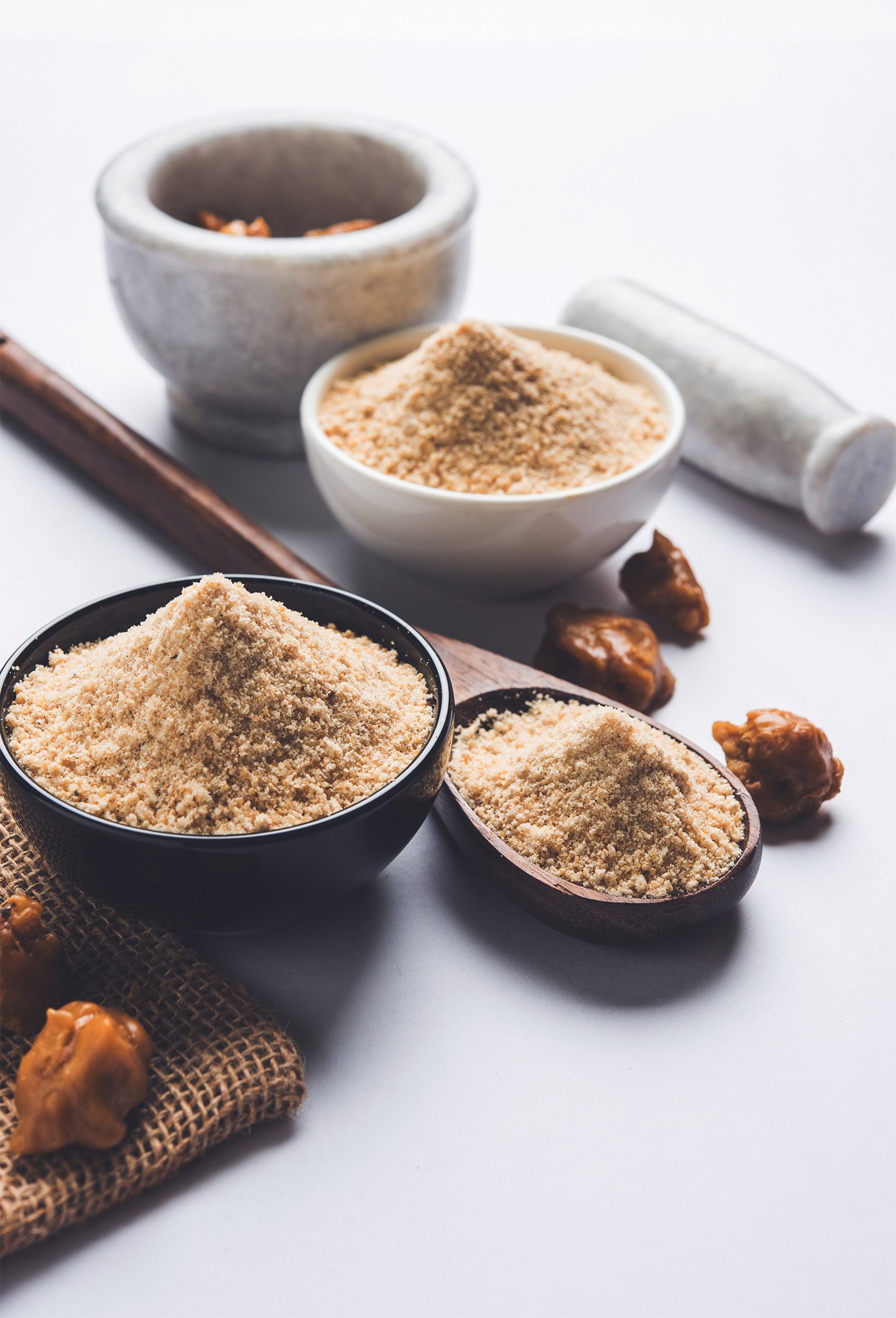 Schalen und Löffel mit gemahlenem Asafoetida, umgeben von getrockneten Harzstücken sowie einem Mörser und Stößel im Hintergrund.