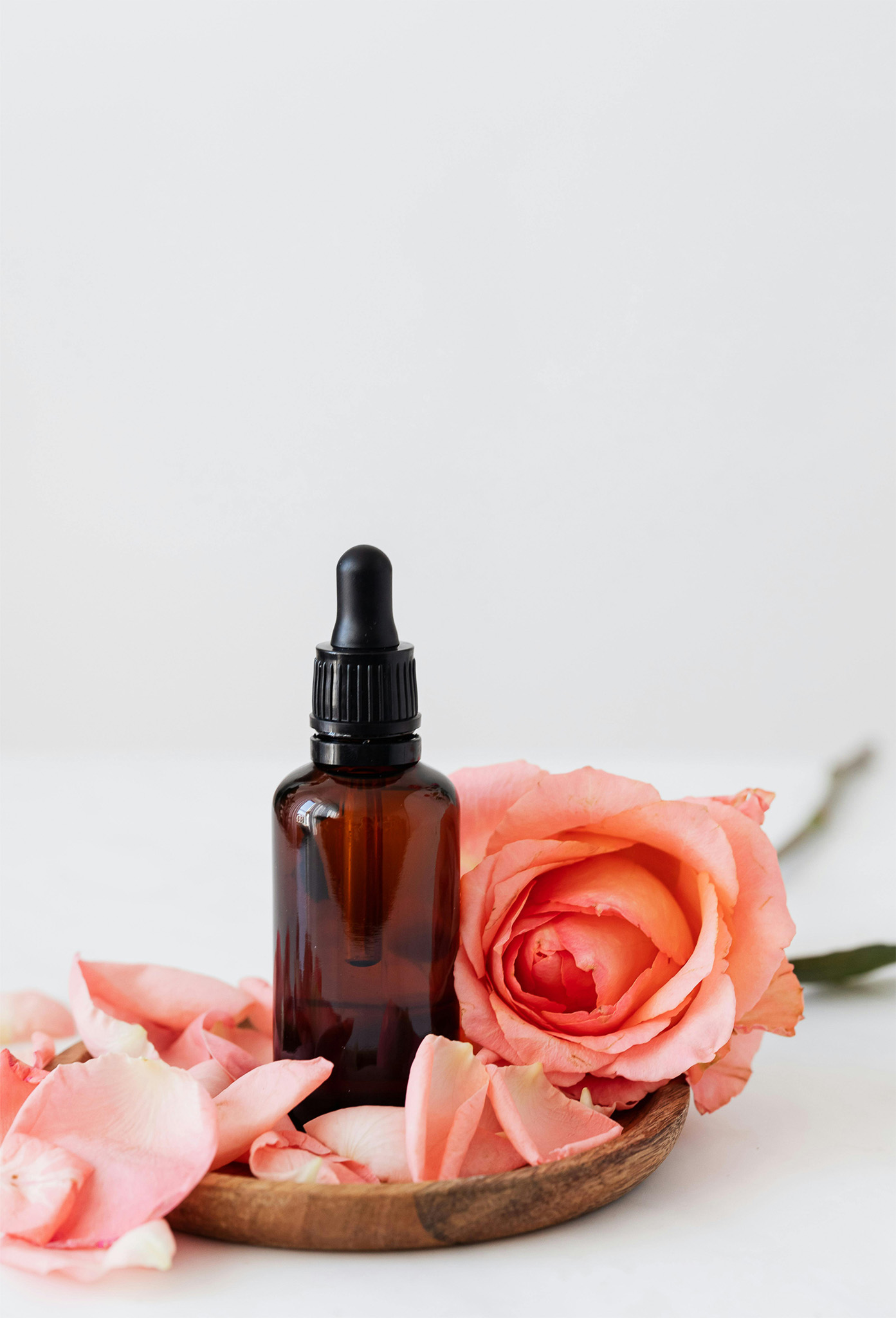Kleine braune Flasche, mit Pippettenaufsatz auf einem kleinen Holzteller. Darum eine Rose und Rosenblüten. 